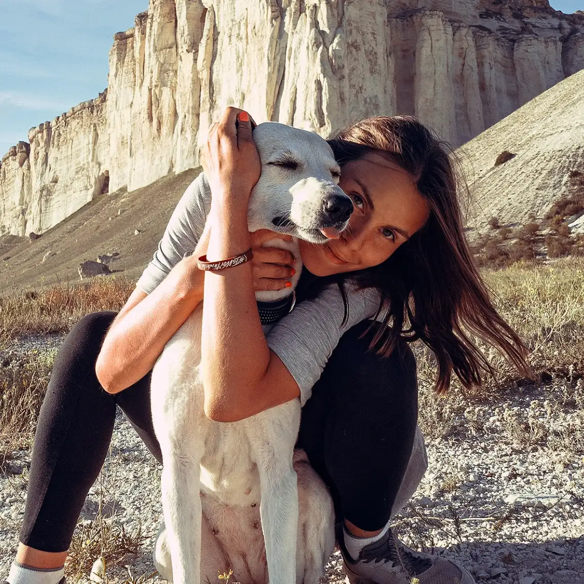 A hiker kneels behind a white Labrador mix hugging them in front of towering cliffs